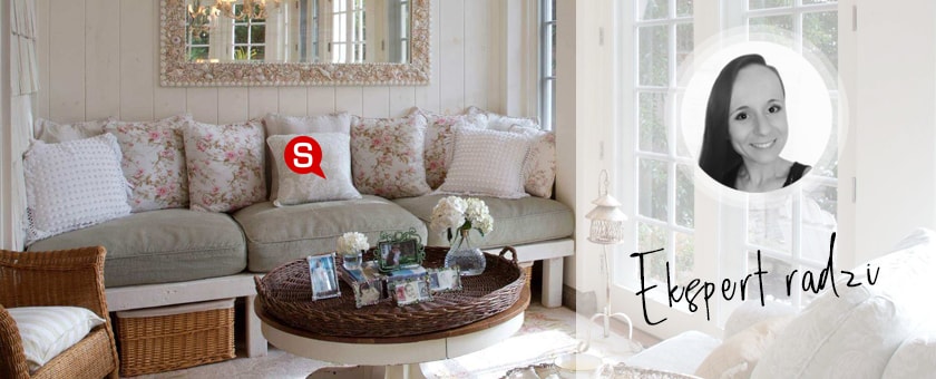 Jasny, przestronny salon z kanapą, nad którą wisi lustro w ozdobnej ramie. Na kanapie znajdują się dekoracyjne poduszki w kwiaty, a całość aranżacji utrzymana jest w odcieniach różu i beżu. Dopełnienie wnętrza stanowią wiklinowe elementy.